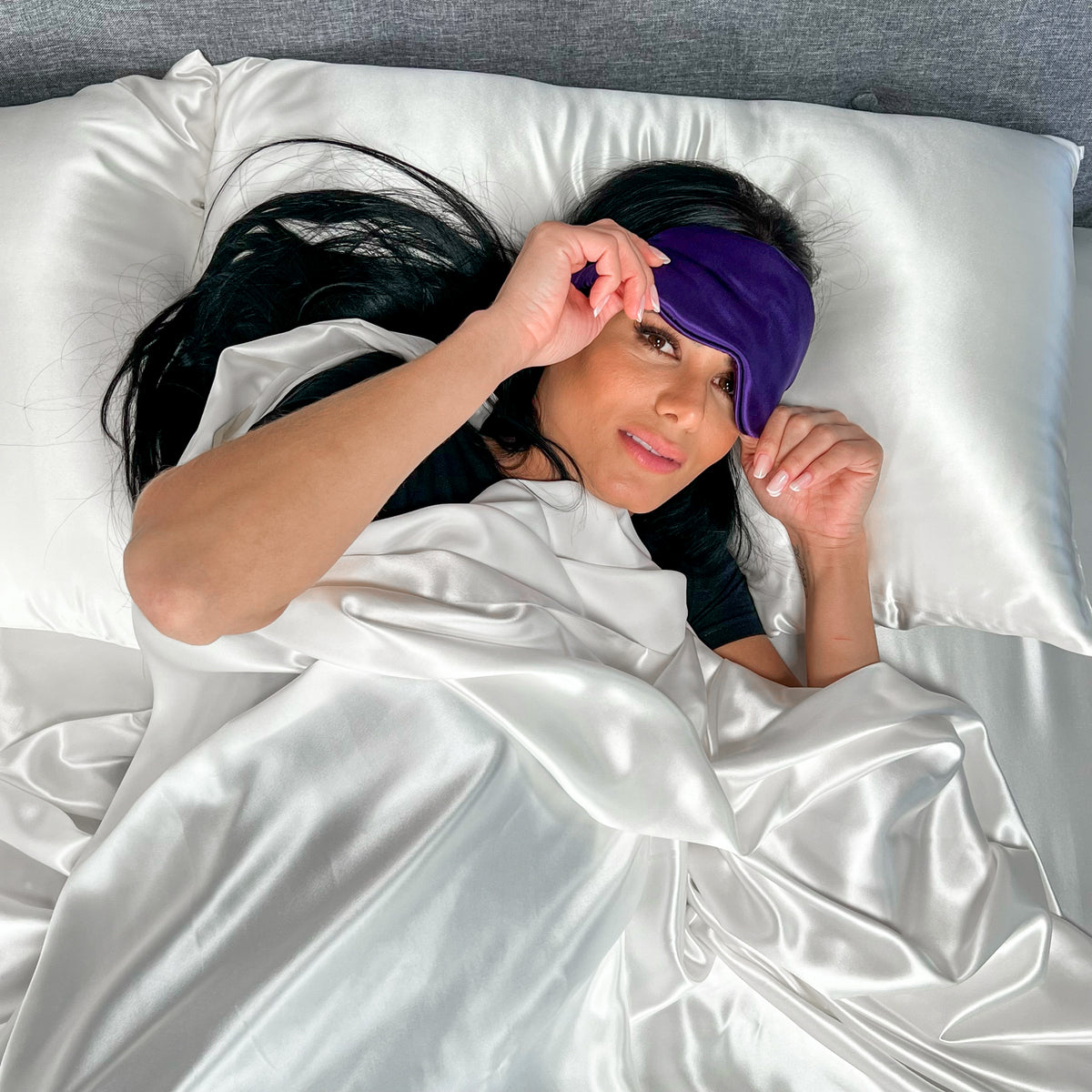 Woman relaxing on a bed with a white silk pillowcase and duvet wearing a plum silk sleep mask