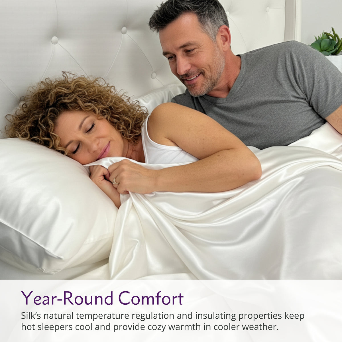 A woman sleeps under a silk blanket while a man lies beside her on a white bed in a bright bedroom.