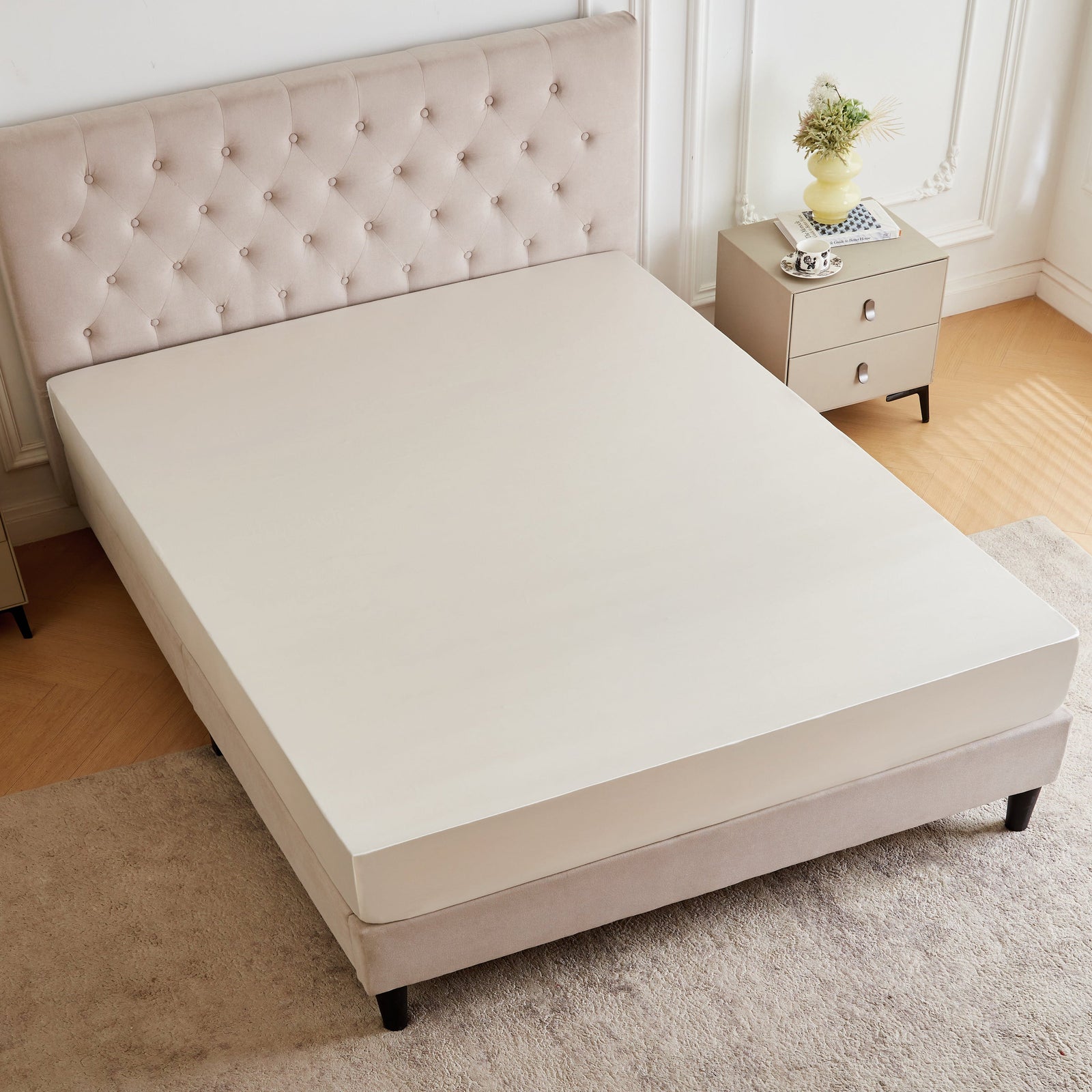 A neatly made bed with a tufted beige headboard, beige mattress, and matching bedside tables with drawers and a yellow vase with flowers, set in a bright, neutral room.
