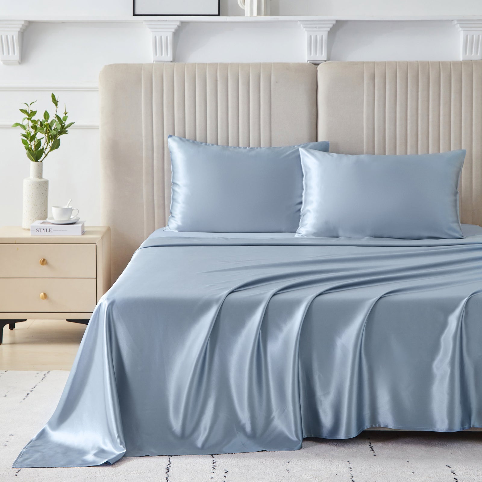 A neatly made bed with light blue satin sheets and two matching pillows, set against a beige upholstered headboard. A nightstand with a plant and books is on the left side of the bed.