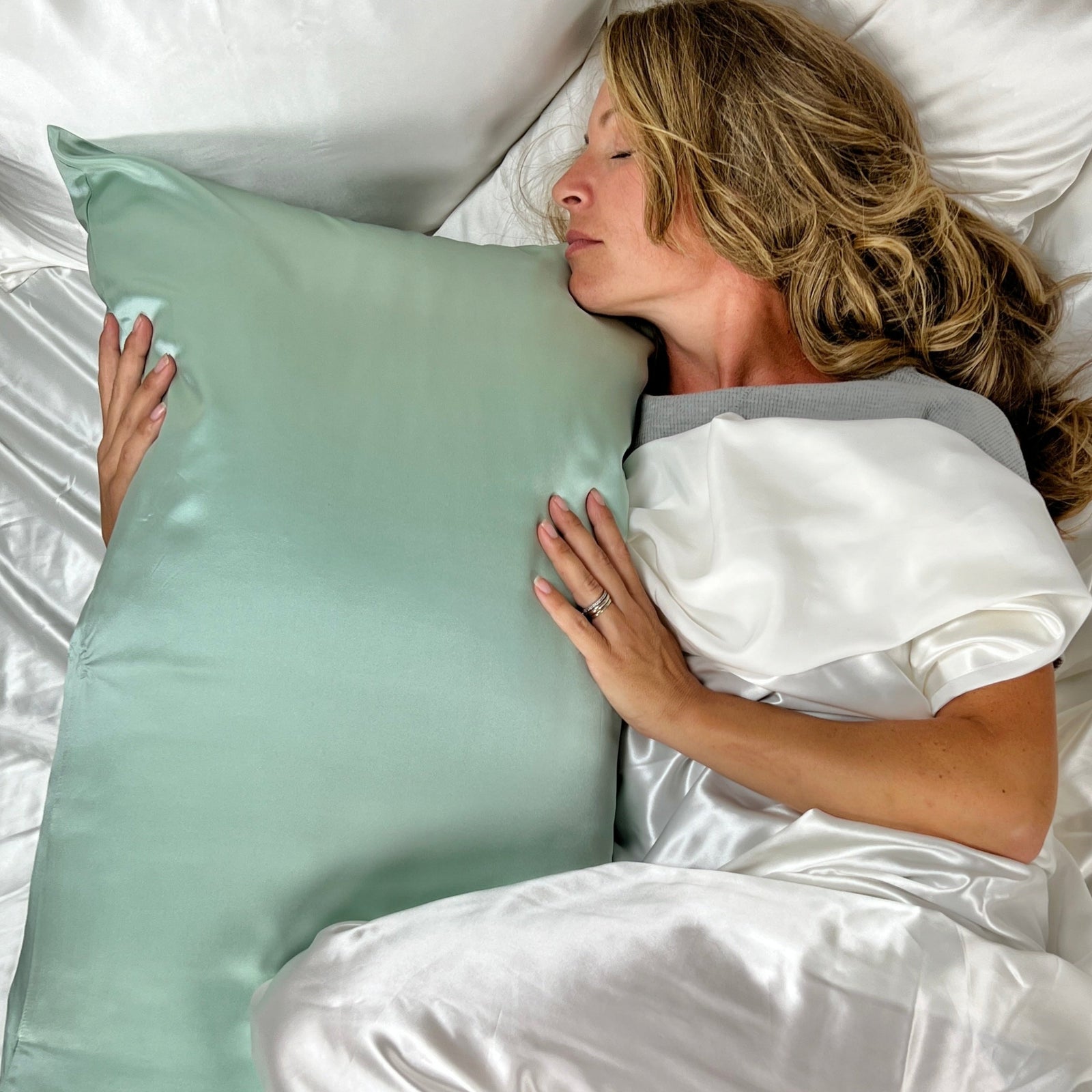 A woman with long blonde hair lies in bed with her eyes closed, hugging a large light green satin pillow. She is covered by white satin sheets and wears a white and gray top.