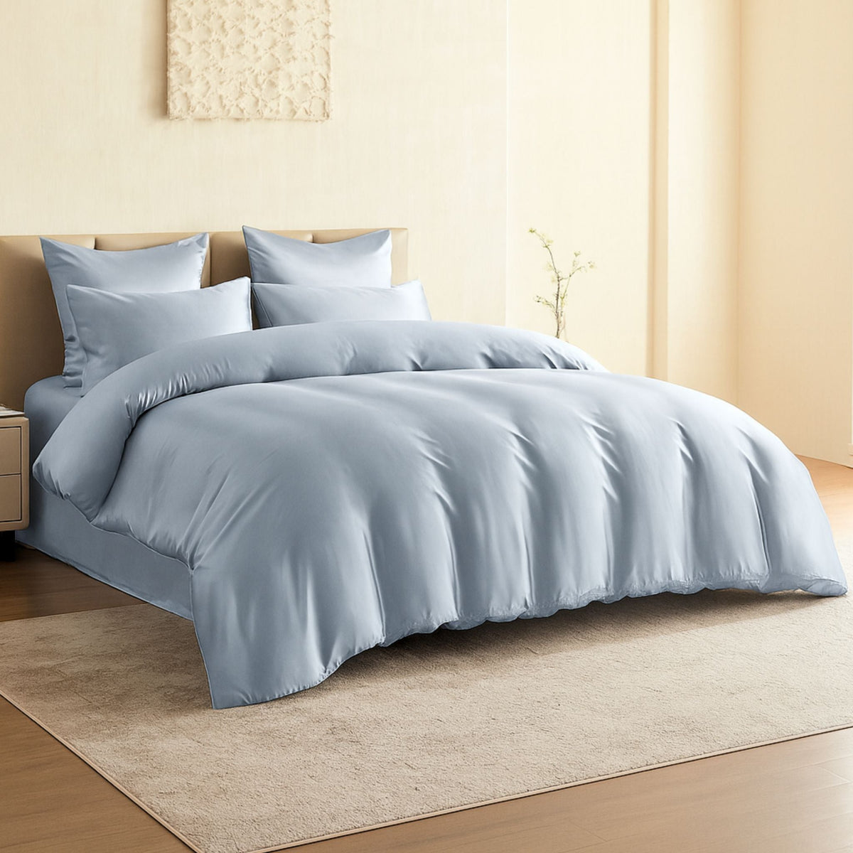 A neatly made bed with light blue bedding and four matching pillows is placed against a beige wall, with a small nightstand and a simple decorative wall piece above the headboard.