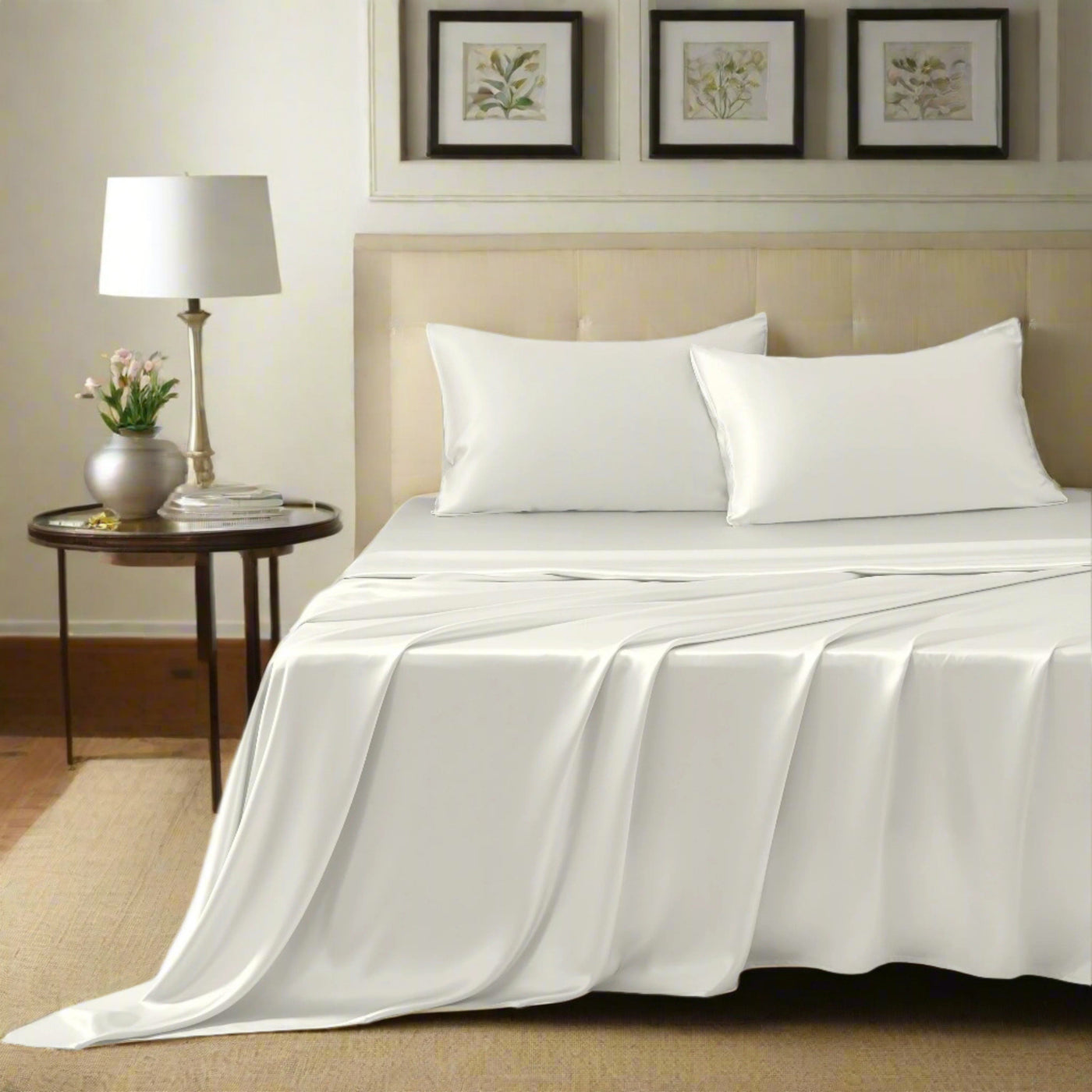 A neatly made bed with a beige tufted headboard, white sheets, and two white pillows. A round wooden nightstand with a lamp and plant is on the left. Three framed botanical prints hang above the bed.