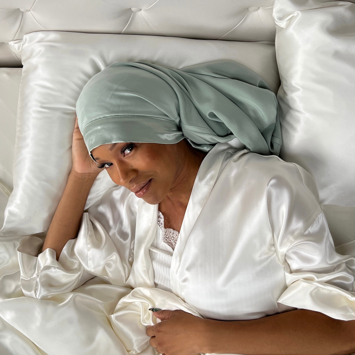 A woman wearing a light green headscarf and a white satin robe lies on a bed with white satin sheets and pillows, resting her head on one hand and looking at the camera.