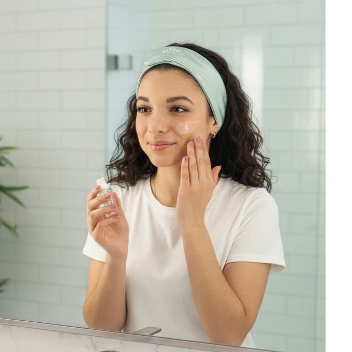 A woman wearing a white t-shirt and a light blue headband applies skincare product to her cheek while looking in a bathroom mirror. Toiletries and a plant are visible in the background.