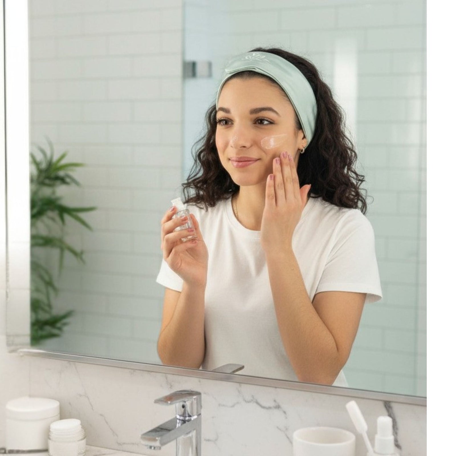 A woman wearing a white t-shirt and a light blue headband applies skincare product to her cheek while looking in a bathroom mirror. Toiletries and a plant are visible in the background.