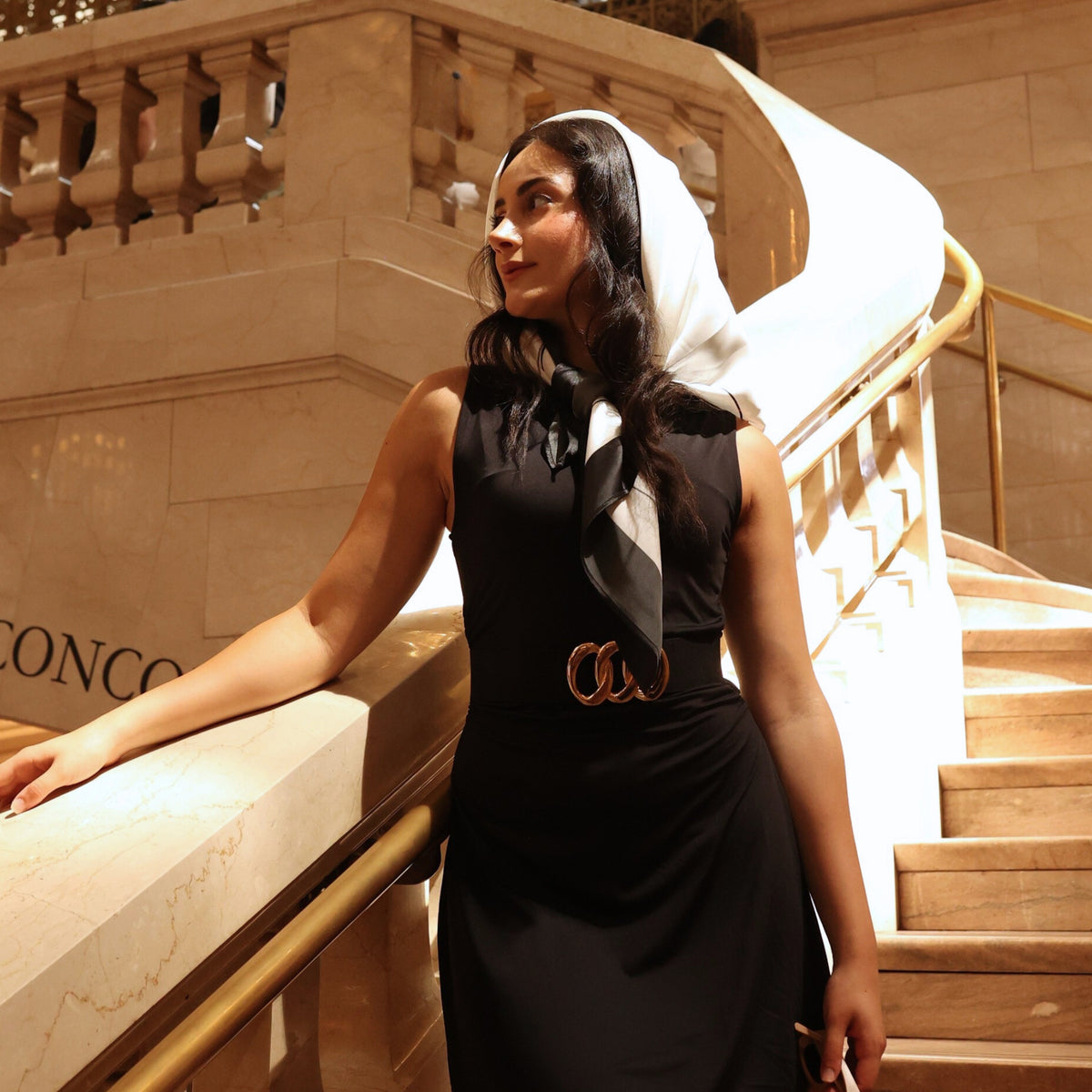 Young woman in a black dress and Silk Square Head Scarf - Black/Ivory from Mulberry Park Silks, standing on a marble staircase, exuding elegance and style.