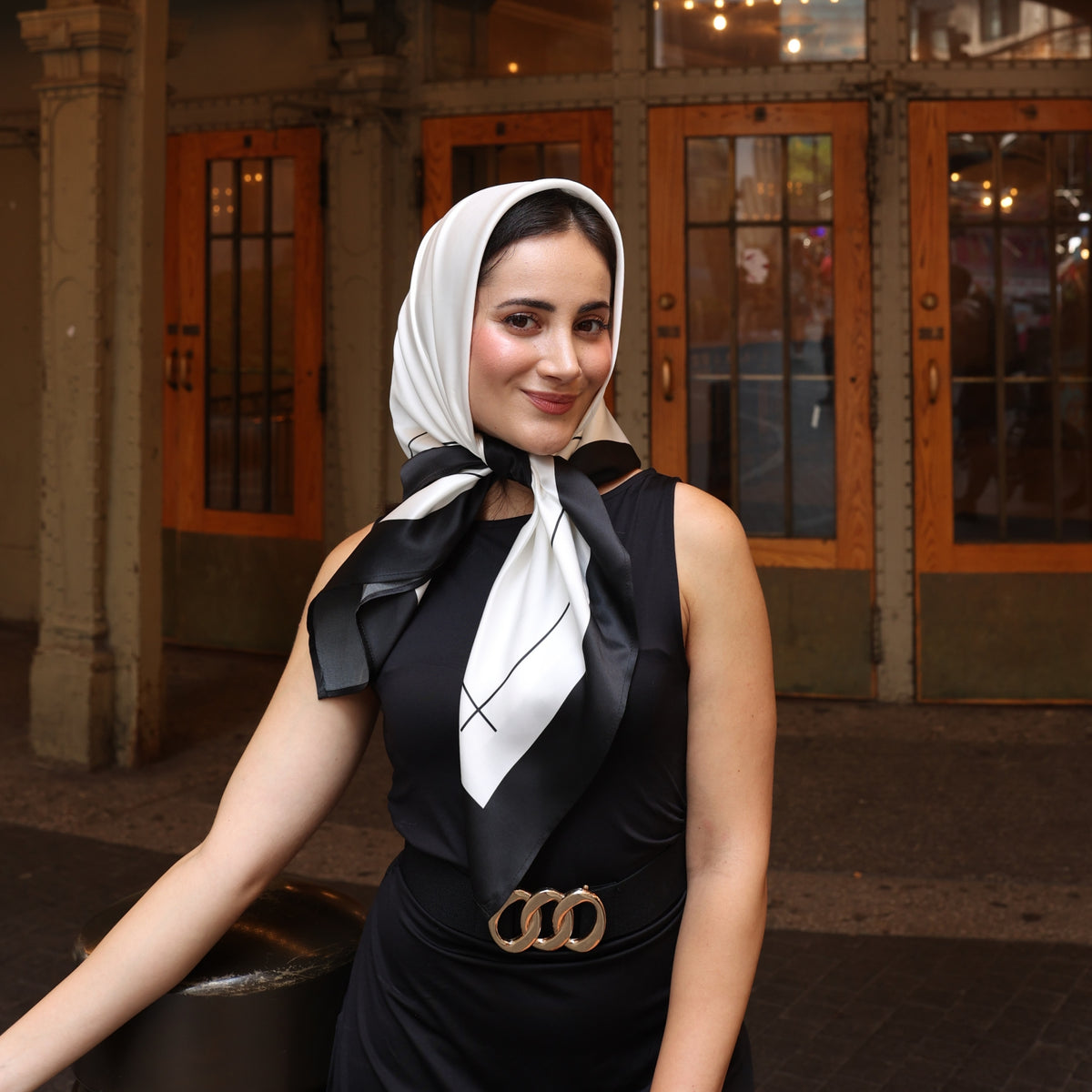 Young woman wearing Mulberry Park Silks Square Head Scarf in Black/Ivory, styled elegantly with a black sleeveless top, smiling outdoors.