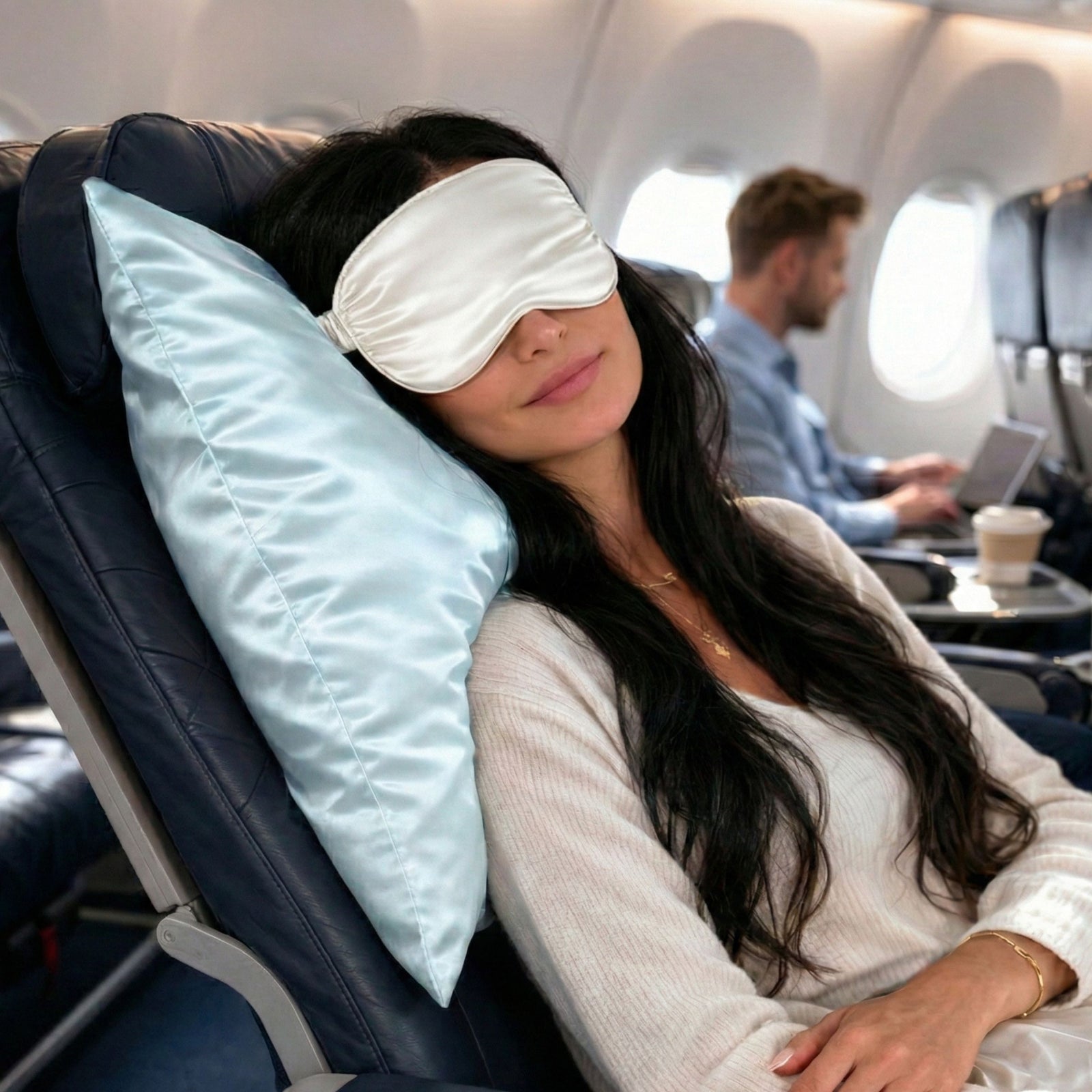 Woman sleeping on an airplane with a blue silk pillowcase and ivory silk sleep mask surrounded by airplane interior.