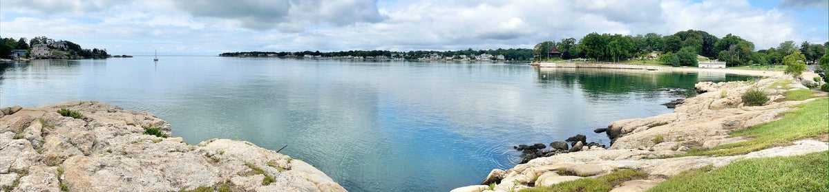 Panoramic view of a calm bay with rocky shore, distant houses, trees, green grass, and a silk-like partly cloudy sky.