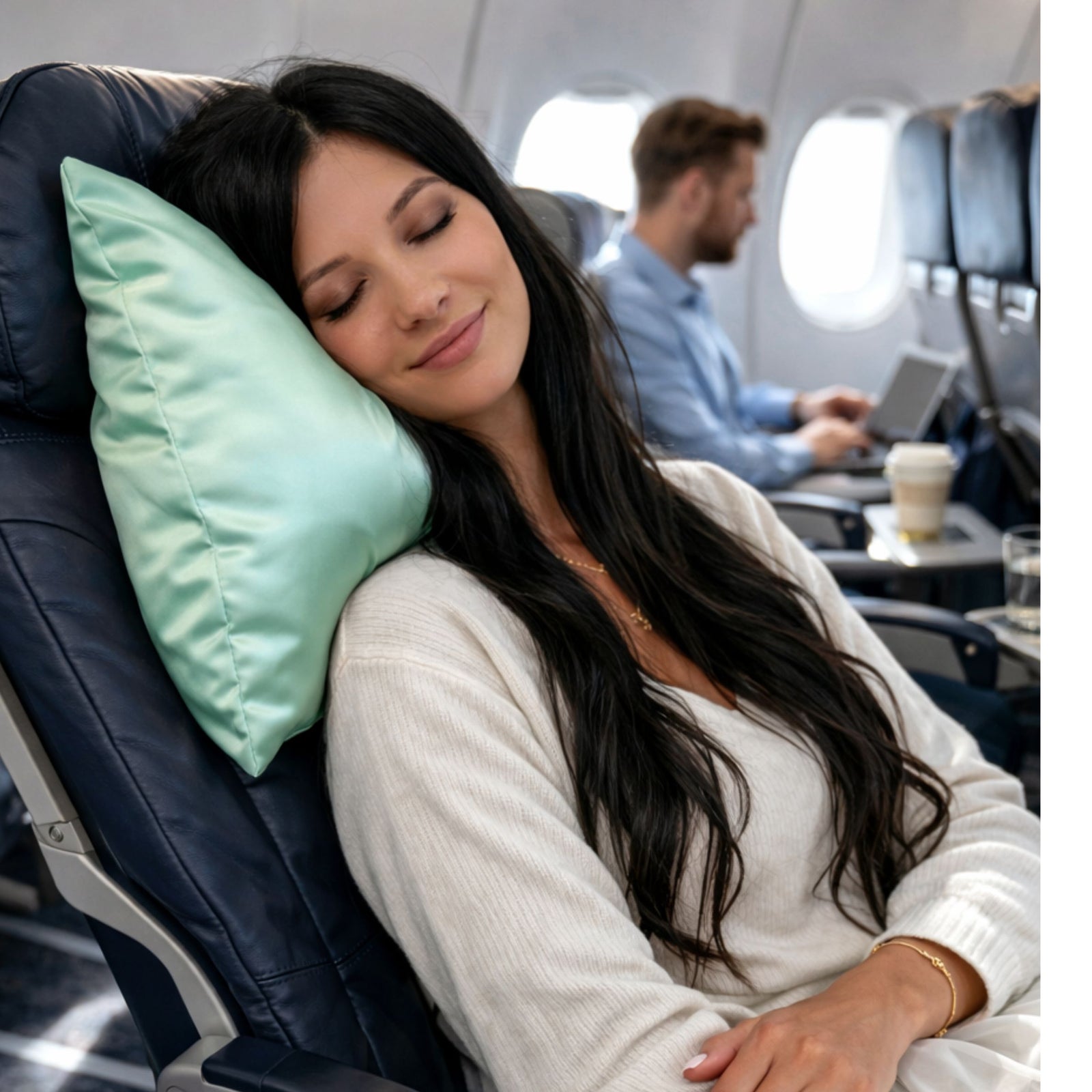 A woman sleeps in her airplane seat with a pure silk travel pillowcase; behind her, a man works on his laptop.