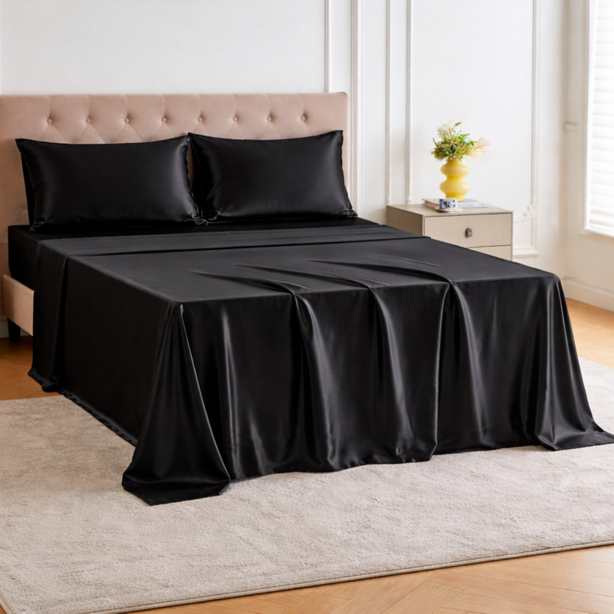 A bed with luxury silk bedding and black satin pillows, beside a nightstand with a yellow vase and flowers in a bright room.