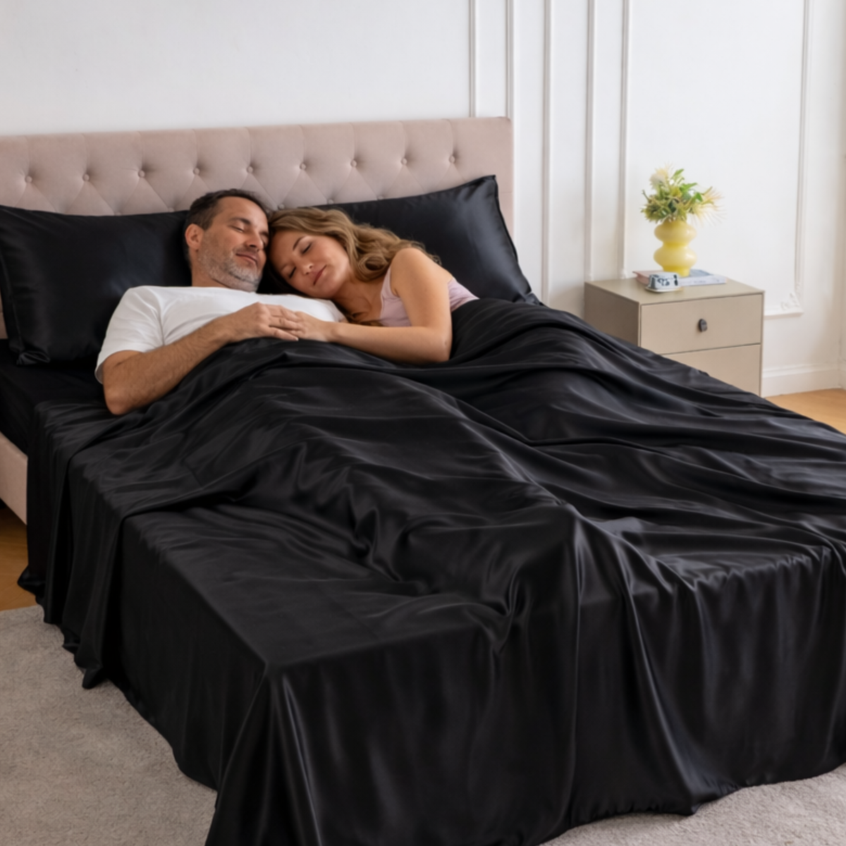 A couple lies in bed under luxury silk bedding, with a nightstand and yellow vase in the background.