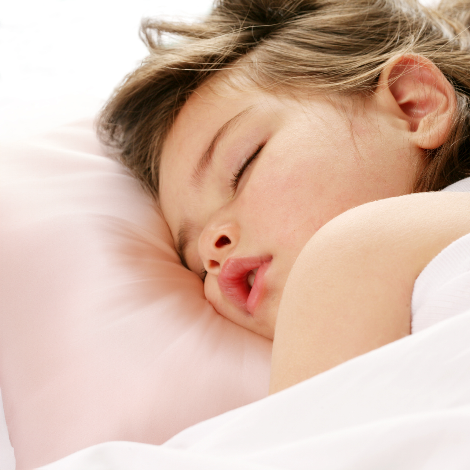 A brown teddy bear sits next to a plain light pink silk pillow on a white background.
