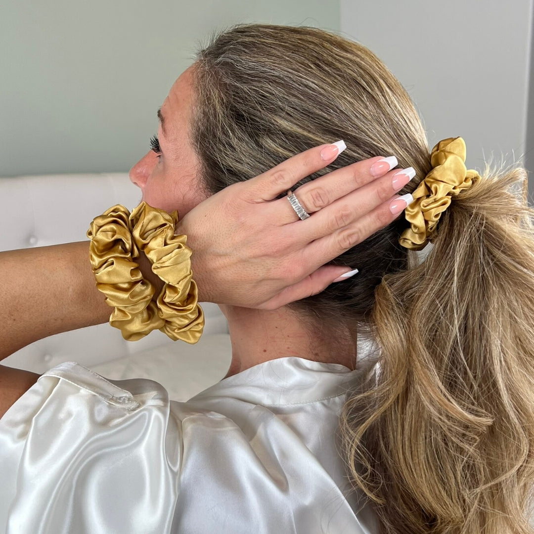 A woman with light brown hair wears gold silk scrunchies in her hair and on her wrist, holding her ponytail.
