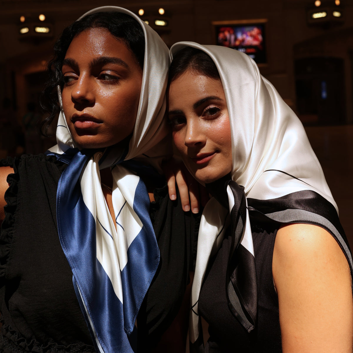 Two women showcase Mulberry Park Silks' Silk Square Head Scarf in Navy/Ivory, elegantly tied under the chin, highlighting the luxurious sheen of 100% Mulberry silk.