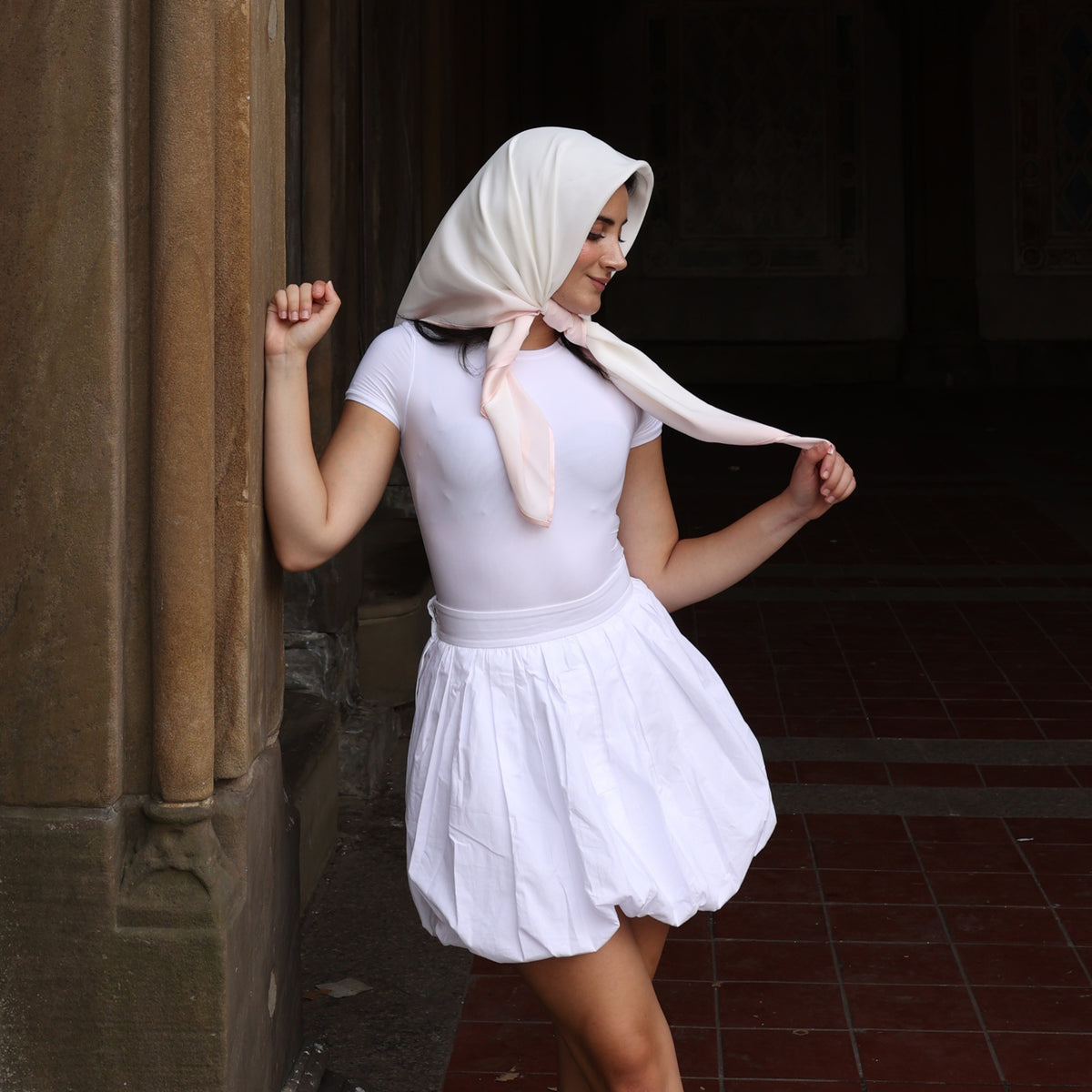 Young woman in a covered arcade wearing a Mulberry Park Silks square head scarf, pink/ivory, styled elegantly with a white outfit, embodying grace and sophistication.