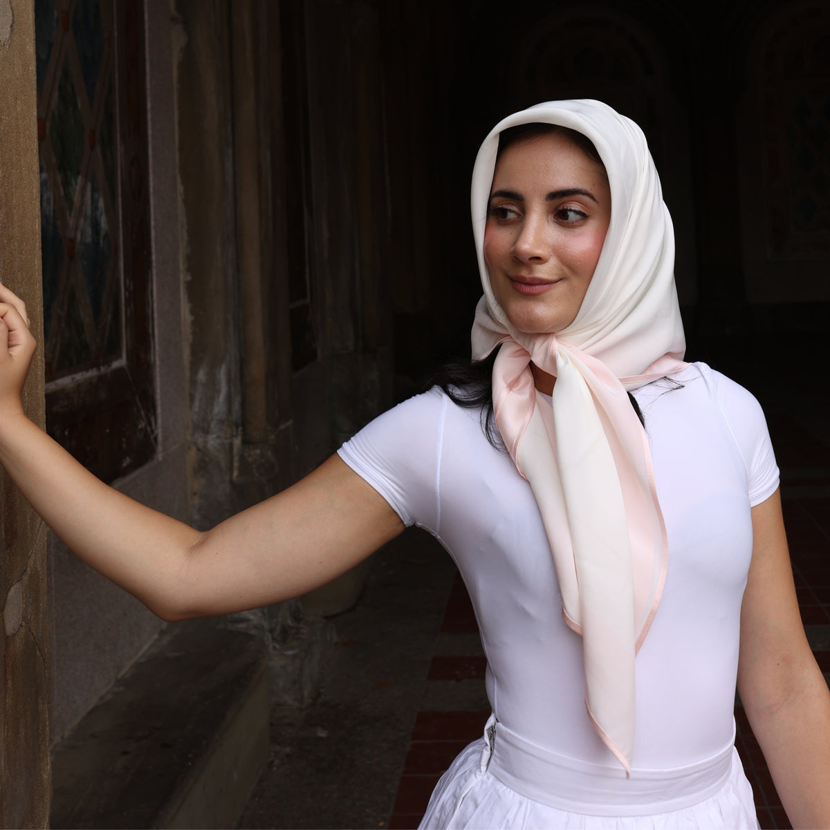 Woman in a stone corridor wearing Mulberry Park Silks' Silk Square Head Scarf - Pink/Ivory, resting her arm on the wall, exuding elegance.