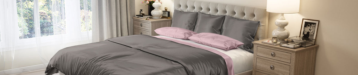 A bedroom with a silk gray and pink bedspread, beige headboard, two nightstands, lamps, and a window with curtains.