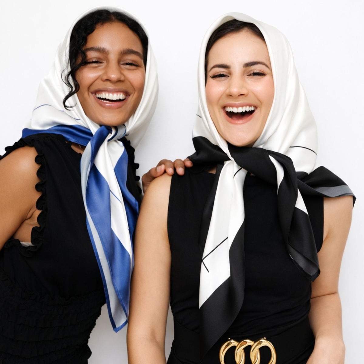 Two women in black outfits smile, each styling a patterned silk scarf on their heads against a plain white background.