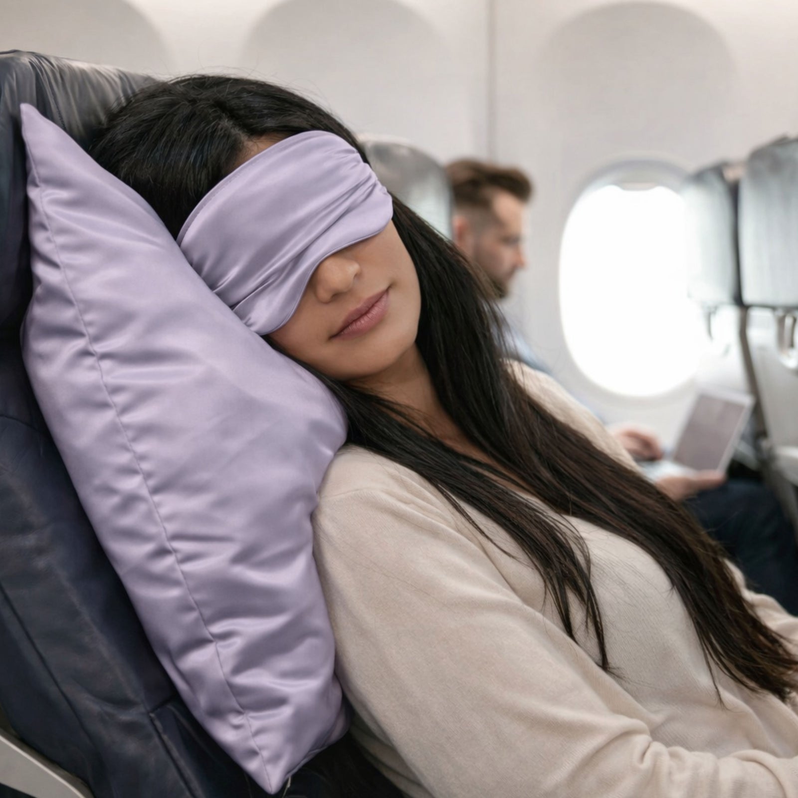 Woman sleeping on an airplane with a lilac travel pillowcase with matching sleep mask, another passenger in the background.