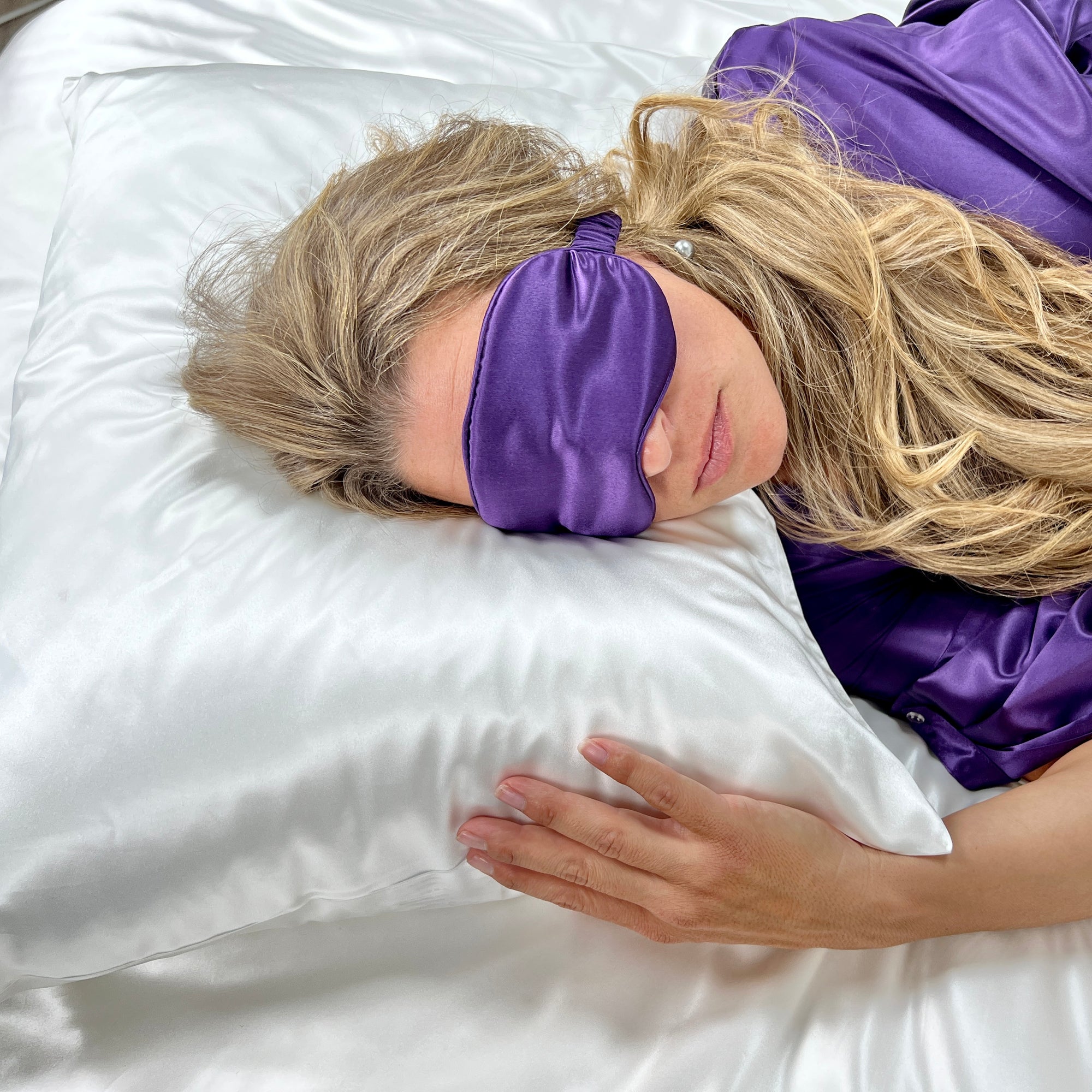 Woman resting on a white silk pillowcase with a matching sleep mask, lying on the bed.