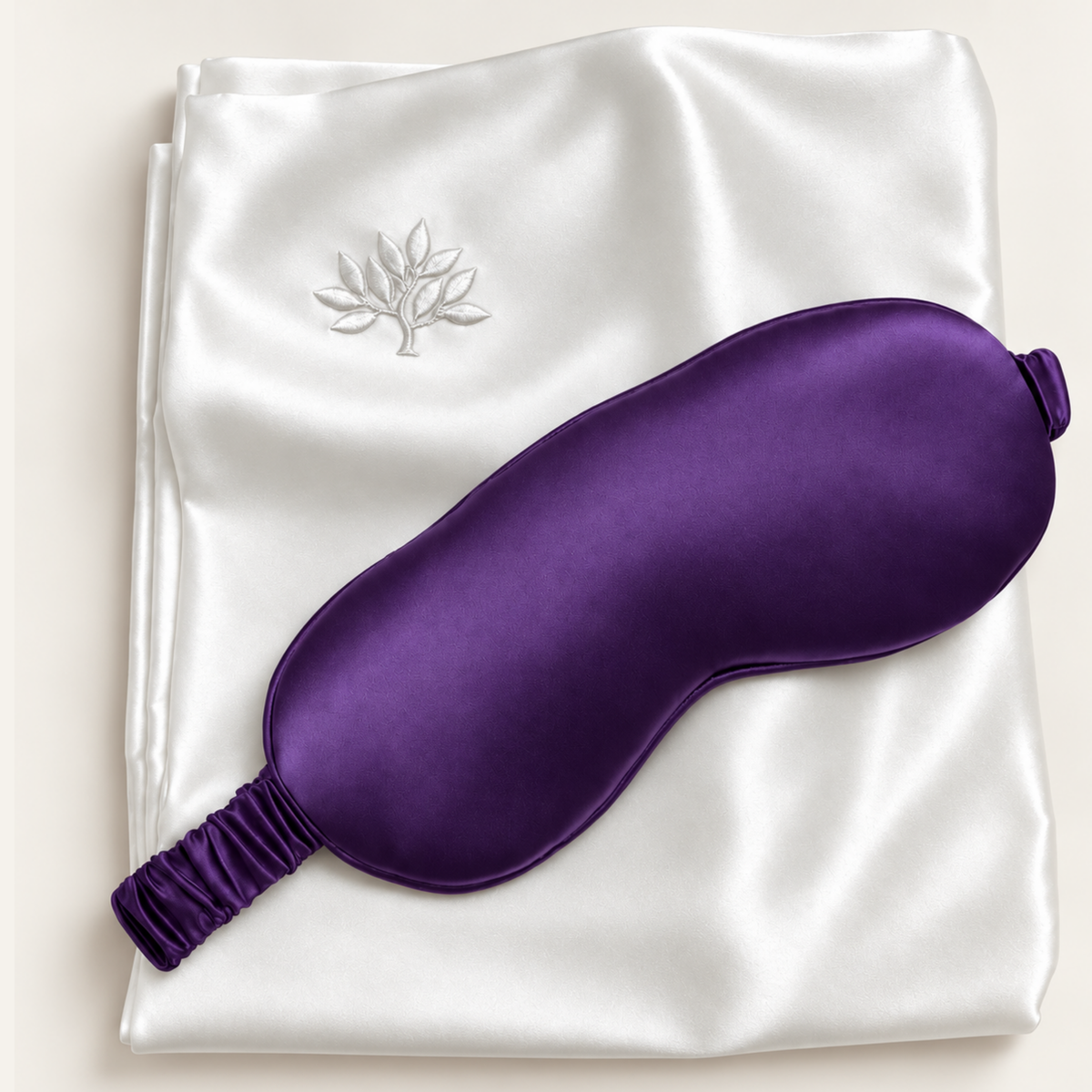 A purple sleep mask rests on folded white mulberry silk with a silver embroidered tree design.