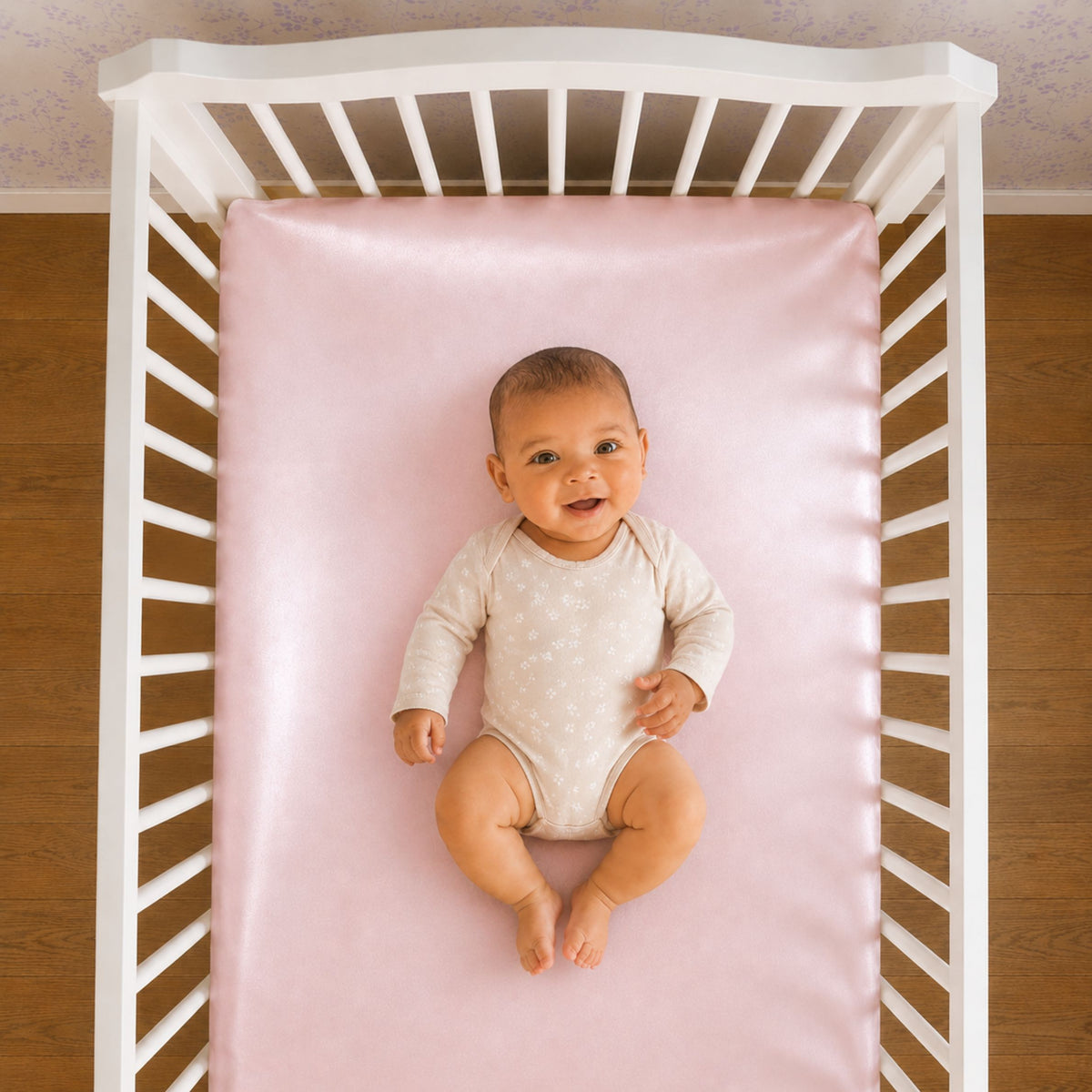 A baby in a light onesie lies on a pink Mulberry silk crib sheet in a white crib, looking up and smiling.