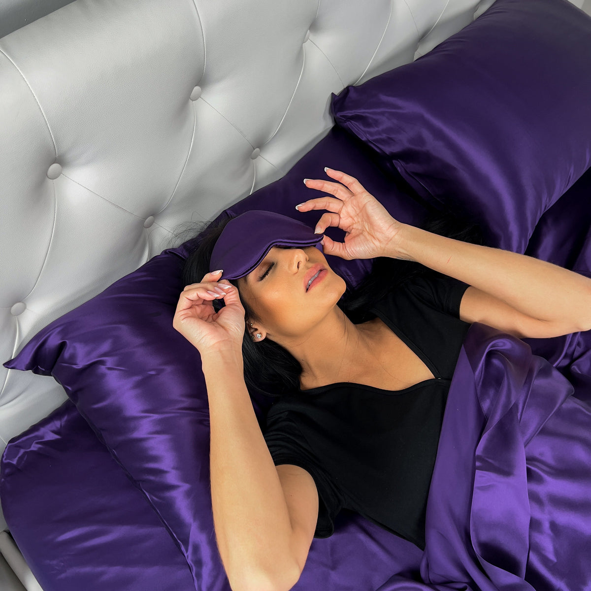 A woman waking up on dark purple silk sheets, slightly lifting her sleep mask off