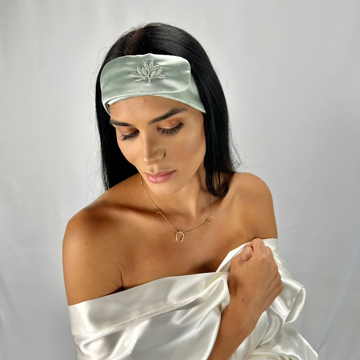 Woman with long dark hair wears a light green silk headband and drapes white satin fabric over her shoulders against a white backdrop.