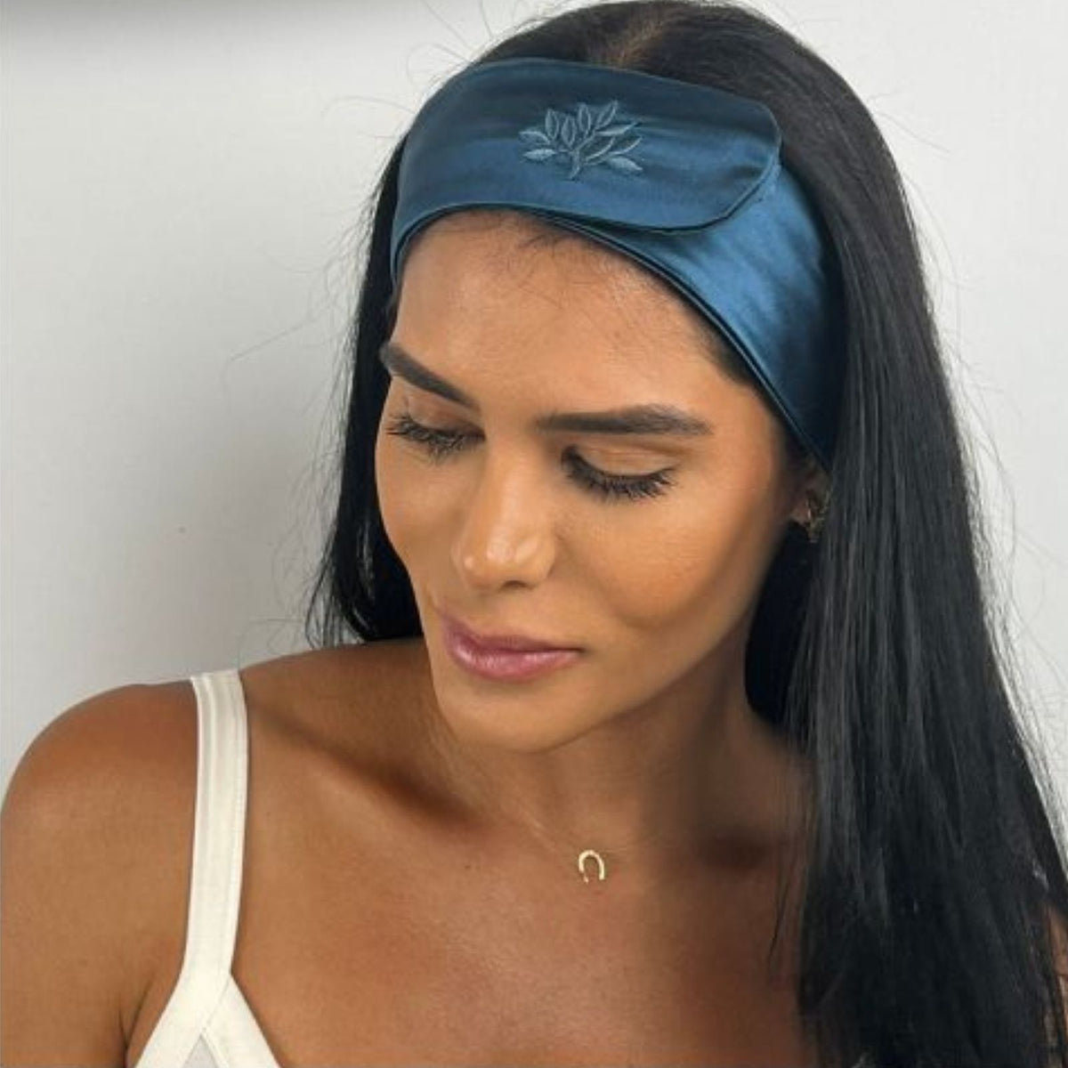 A woman with long dark hair wears a blue silk headband and a white top, looking downward against a plain background.